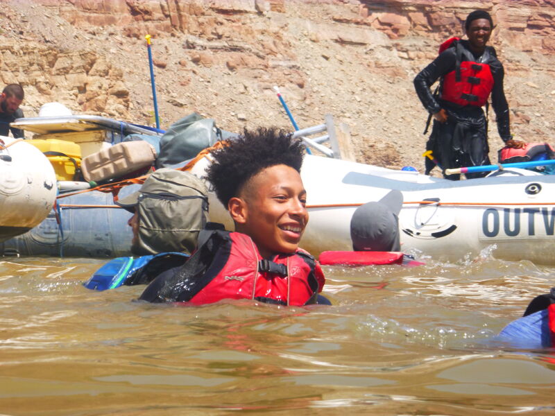 The image shows a group of people enjoying a rafting trip. A young man is in the foreground, swimming in the water with a life jacket on. In the background, there's a raft with other people, some wearing life jackets as well. The raft has the word "OUTW" printed on its side. The background features a rocky, desert-like landscape. It seems like a sunny day, and everyone is having a good time on the river.